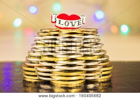 Pyramid of coins with the inscription love on top. Blurry bright background with lights on a dark table