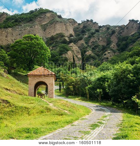 Mtskheta, Georgia. View Of Scenic Area Of Shiomgvime Or Shio-Mgvime Monastery With Small Brick Arched Construction, The Entrance To The Underground Cave And Winding Paved Walkway In Sunny Spring Day.