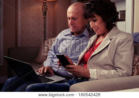A mid shot of man surfing the web and riant woman regarding her phone. A pair sitting on the brown settee with patterned pillows