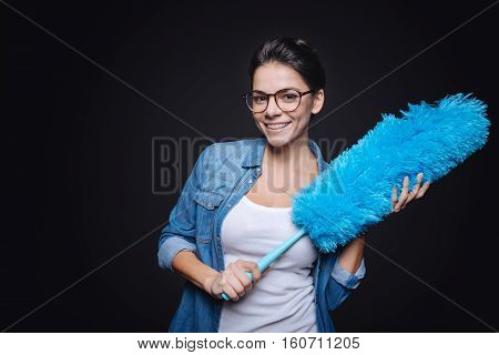 Happy from cleaning. Delighted cheerful happy woman holding a dusting brush while standing against black background and smiling