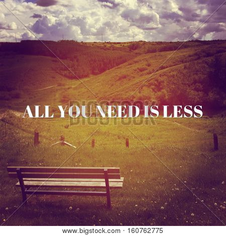 Inspirational quote on park landscape with empty bench overlooking meadow in autumn with hills,forest trees and cloudy sky background. Grass, fence and wildflowers in foreground. All you need is less.  Lighting effects.