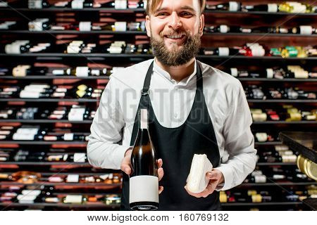 Portrait of a sommelier in uniform with brie cheese and bottle of wine at the restaurant or supermarket. Choosing wine according to the type of cheese. Bottle with empty label to copy paste
