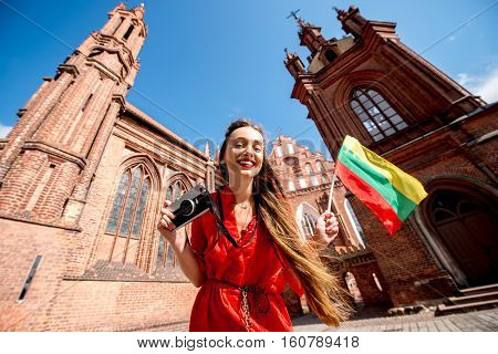 Young female tourist with photo camera and lithuanian flag standing in front of the famous gothic church in the old town of Vilnius. Woman having great vacations in Lithuania