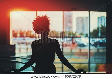 Silhouette of young black curly Brazilian girl in front of window standing in supermarket with blurred escalator behind her and cityscape out of the window Rio de Janeiro Brazil