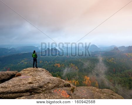 Hiker In Green Windcheater, Cap And Dark Trekking Trousers Stand On Mountain Peak Rock.