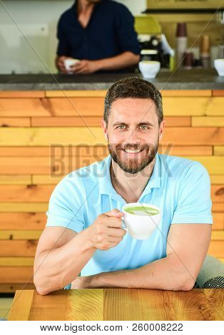 Man Bearded Guy Drinks Cappuccino At Wooden Table Cafe. Cafe Visitor Happy Face Enjoy Coffee Caffein