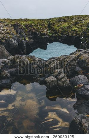 Close-up Shot Of Mossy Rocky Cliff In Shape Of Arch With Blue Ocean On Background In Arnarstapi, Ice