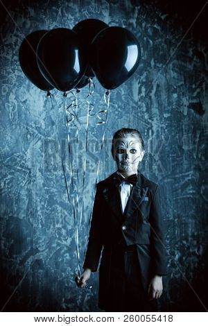 Portrait of a child boy with carnival make-up of a skeleton wearing elegant tail-coat. Studio shot over dark grunge background. Halloween. Dia de los muertos. The Day of the Dead.