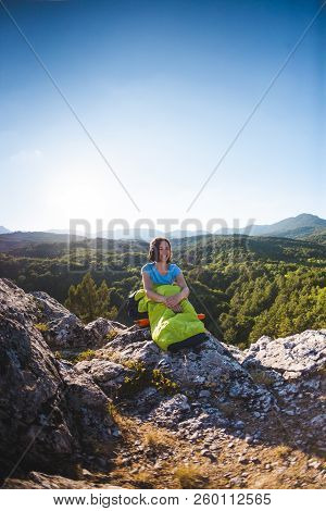 A Woman Is Sitting In A Sleeping Bag At Sunset. Camping On Top Of A Mountain. A Smiling Girl Rests A