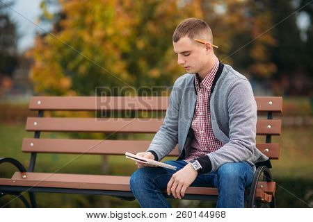A Cute Student Sits On The Bench And Writes Down His Thoughts In His Notebool Using A Pensil. Study 