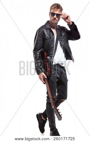 attractive guitarist in black leather jacket walks to side on white background and fixes his sunglasses, full body picture