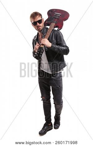 fashion man with guitar on shoulder walking on white background while holding leather jacket's collar, full body picture