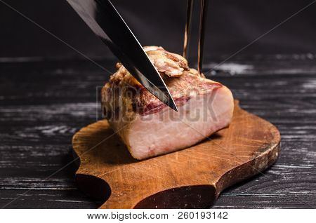 The piece of Meat is cutting using a carving knife and carving fork. A piece of smoked bacon is cutting on a wooden cutting board.
