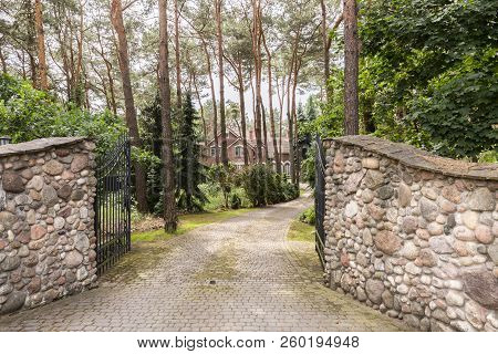 Metal Gate Open To A Cobblestone Driveway To A Luxurious Summer Villa With Brick Facade In The Fores