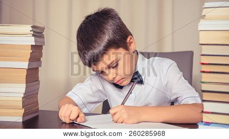 Schoolboy Does Homework At The Table, Next To Two Stacks Of Books. Chilhood, Education Concept. Tone
