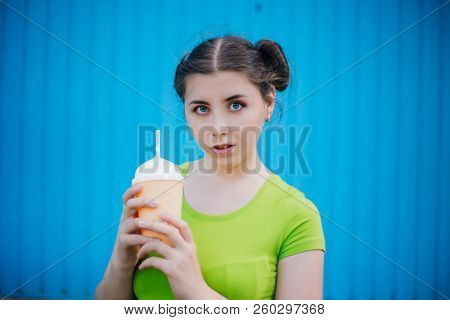 Portrait Of A Girl Against A Bright Blue Wall. Emotional Girl Holding A Plastic Glass With A Refresh