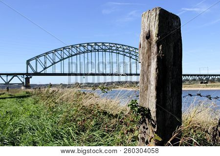 Railway Bridge Spanning The River Rhine Near Arnhem And Oosterbeek, With A Wooden Landmark In The Fr