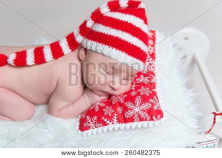 Charming infant baby in striped bonnet sleeping on red pillow on small wooden sledge crib with plush Christmas cane toy nerby