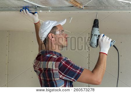Young Man In Goggles Fixing Drywall Suspended Ceiling To Metal Frame Using Electrical Screwdriver On