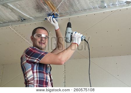Young Man In Goggles Fixing Drywall Suspended Ceiling To Metal Frame Using Electrical Screwdriver On