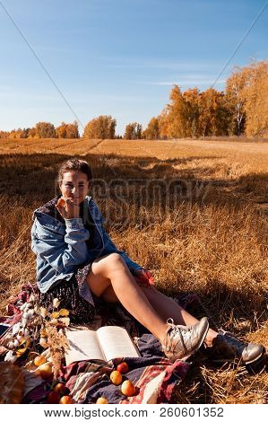 Picnic In The Open Air: A Young Woman In A Jeans Jacket And A Dress Enjoys Nature, Sitting On A Plai