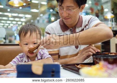 Asian Father Feeding Food To Cute Little Asian 2 Years Old Toddler Boy Child While Watching Smartpho