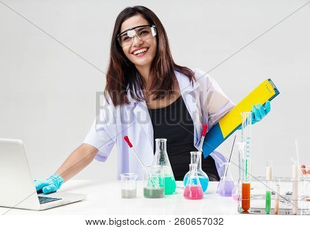 The Chemist In Protective Eyeglasses Working By Test Tube With Reagent In Lab,she Is Using Laptop To