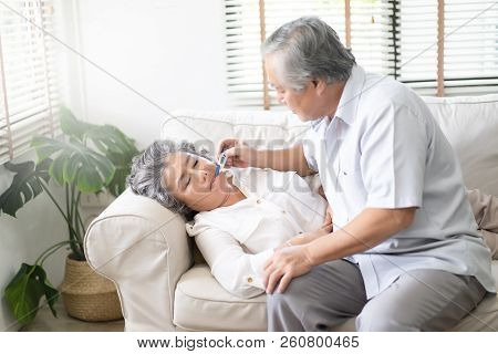 Sick Old Woman With Thermometer Laying In Sofa And Husband Hand Taking Temperature. Husband Checking