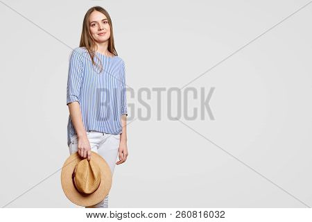 Attractive Relaxed Young European Woman Wears Fashionable Striped Blouse, White Trouses, Holds Straw