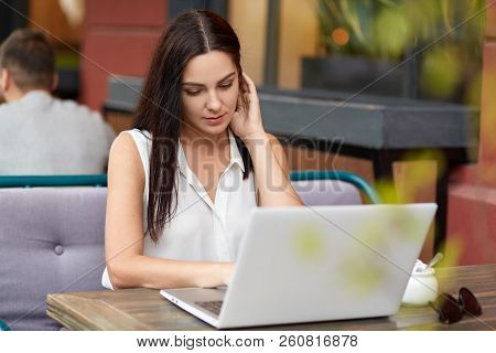 Photo Fo Concetrated Female Freelancer Watches Tutorial Online, Uses Laptop Computer For Work, Sits 