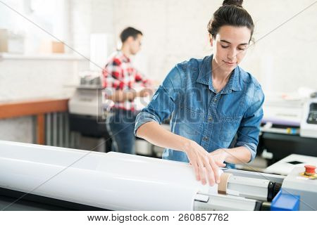 Serious Confident Young Woman  With Hair Bun Loading Wide Format Printer While Working At Printing H