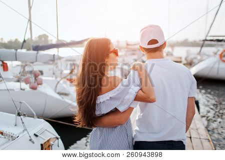Couple Stands And Look On Yachts That Are On Each Sode From Pier. They Hug Each Other. Girl Wears Dr