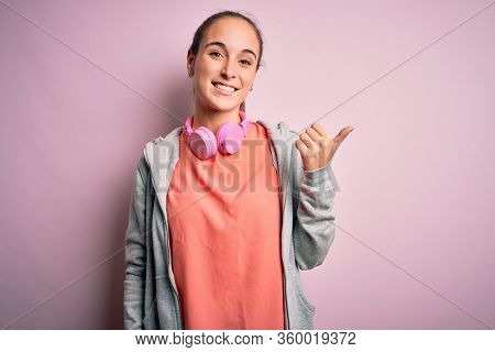 Beautiful sporty woman doing sport listening to music using headphones over pink background smiling with happy face looking and pointing to the side with thumb up.