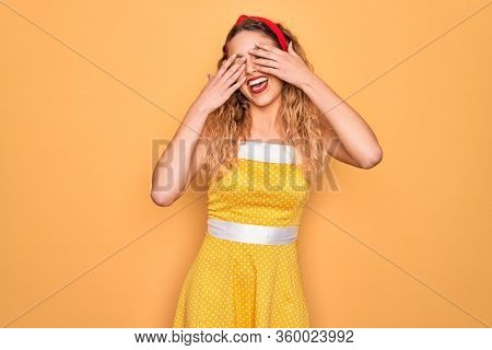 Beautiful blonde pin-up woman with blue eyes wearing diadem standing over yellow background covering eyes with hands smiling cheerful and funny. Blind concept.
