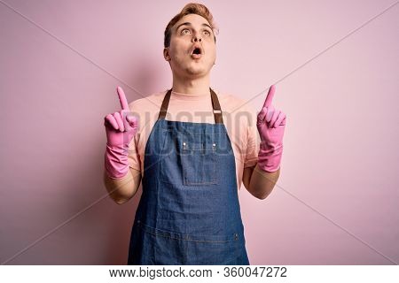 Young handsome redhead cleaner man doing housework wearing apron and gloves amazed and surprised looking up and pointing with fingers and raised arms.
