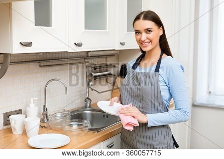 Beautiful Young Woman In Apron Drying The Dishes, Wiping Them Off With A Towel, Looking At Camera An