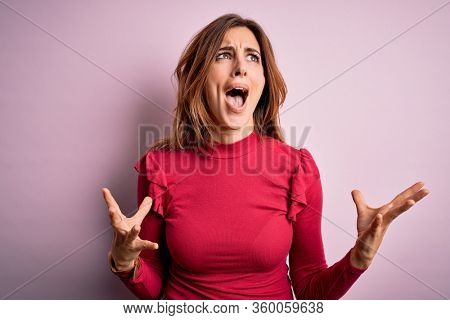 Young beautiful brunette woman wearing casual sweater stnding over pink background crazy and mad shouting and yelling with aggressive expression and arms raised. Frustration concept.