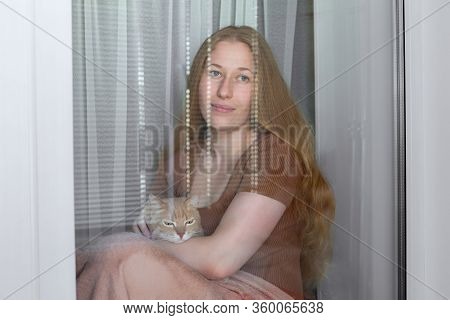 The Girl Is Sitting On A Window Sill; Wrapped In A Blanket And Holding A Cat In The Hands Of A Coron