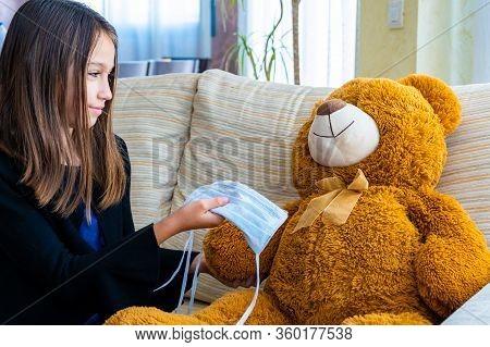 Beautiful Little Girl In A Black Jacket Playing With Her Brown Teddy Bear Who Has Put A Mask To Prot