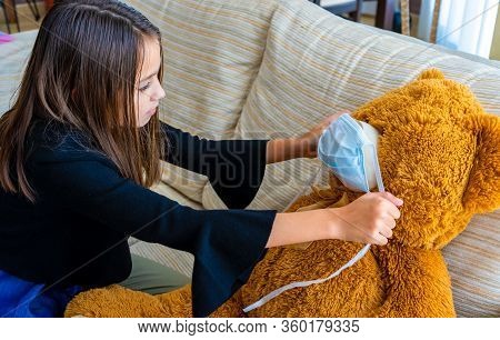 Beautiful Little Girl In A Black Jacket Playing With Her Brown Teddy Bear Who Has Put A Mask To Prot