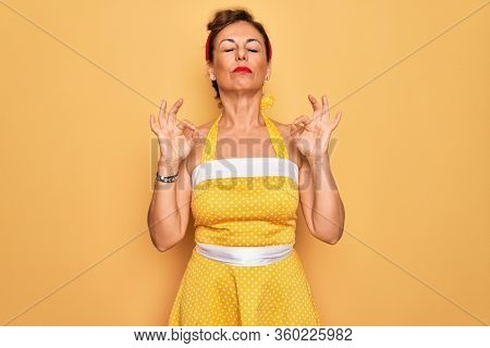 Middle age senior pin up woman wearing 50s style retro dress over yellow background relaxed and smiling with eyes closed doing meditation gesture with fingers. Yoga concept.