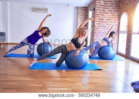 Young beautiful group of sportswomen smiling happy practicing yoga. Stretching using fitness ball at gym
