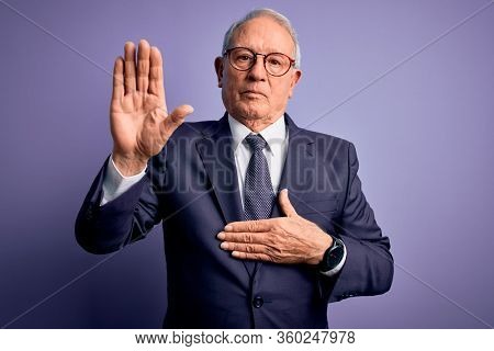 Grey haired senior business man wearing glasses and elegant suit and tie over purple background Swearing with hand on chest and open palm, making a loyalty promise oath