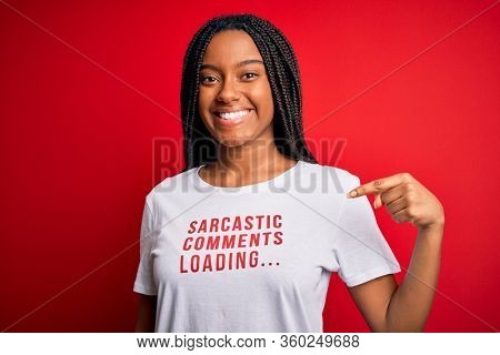 Young african american woman wearing sarcasm coments text on t-shirt over red background with surprise face pointing finger to himself