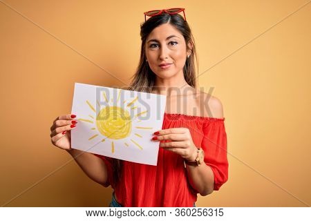 Young beautiful brunette woman holding paper with sun draw over isolated yellow background with a confident expression on smart face thinking serious