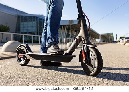 Legs Of A Man Standing On E-scooter Parked On Sidewalk At Cityscape On The Sunset. Trendy Urban Tran