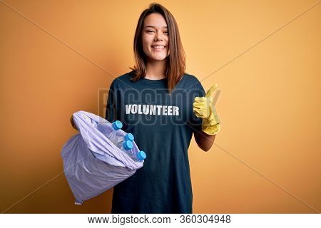 Beautiful volunteer girl caring environment doing volunteering holding bag with rubish bottles happy with big smile doing ok sign, thumb up with fingers, excellent sign