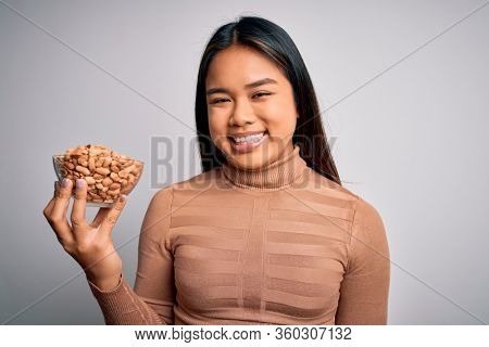 Young asian girl holding bowl of healthy peanuts eating as snack for diet over white background with a happy face standing and smiling with a confident smile showing teeth