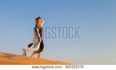 A Woman In A Black Dress With A Cloth In Her Hands In A Strong Wind Alone In The Desert. A Girl Walk