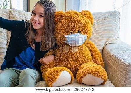 Beautiful Little Girl In A Black Jacket Playing With Her Brown Teddy Bear Who Has Put A Mask To Prot
